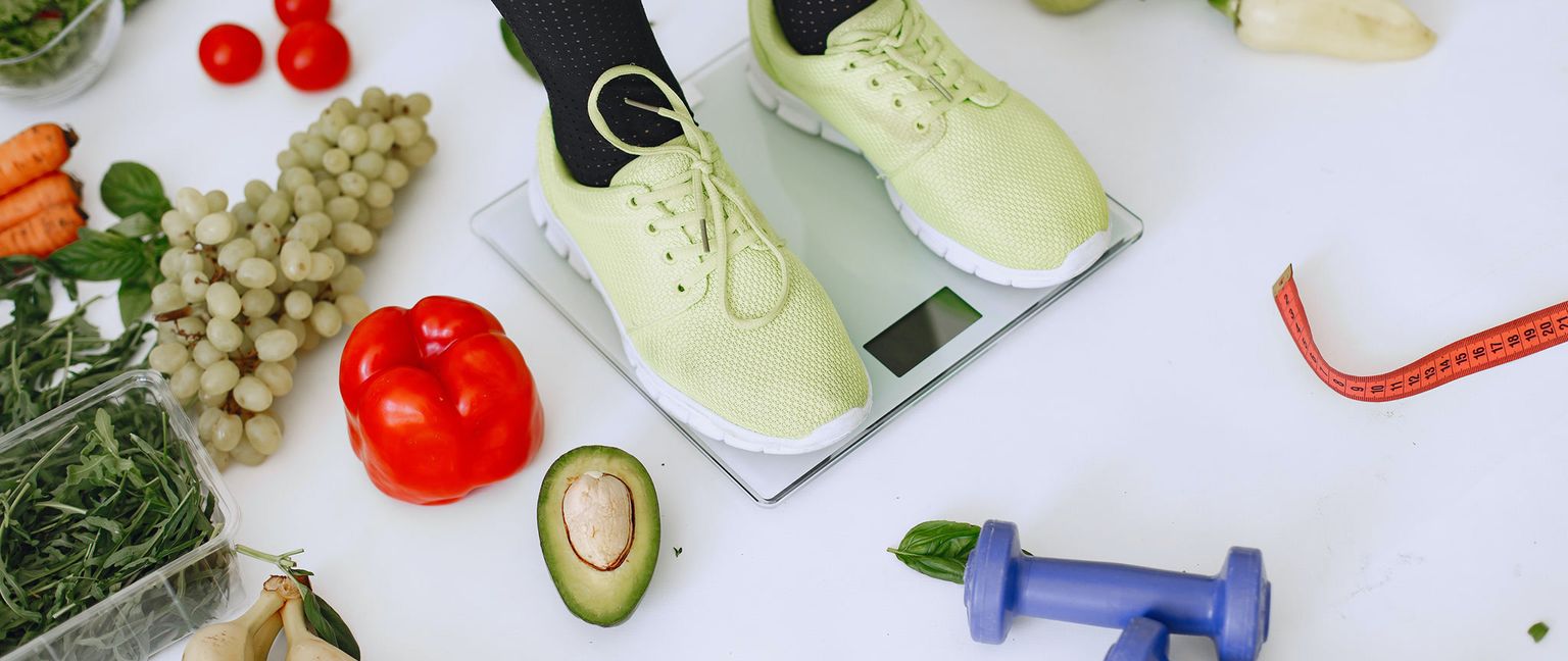 A pair of lime green athletic shoes on a scale are surrounded by various healthy foods including grapes, carrots, a red pepper, avocado, bananas, and greens. A red measuring tape and a blue hand weight are also visible.