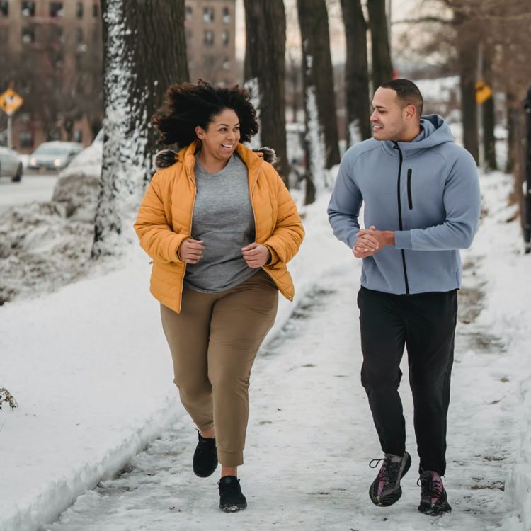 A couple running on a snowy sidewalk, smiling and looking at each other. The woman is wearing a yellow puffer jacket and the man is wearing a gray hooded jacket.