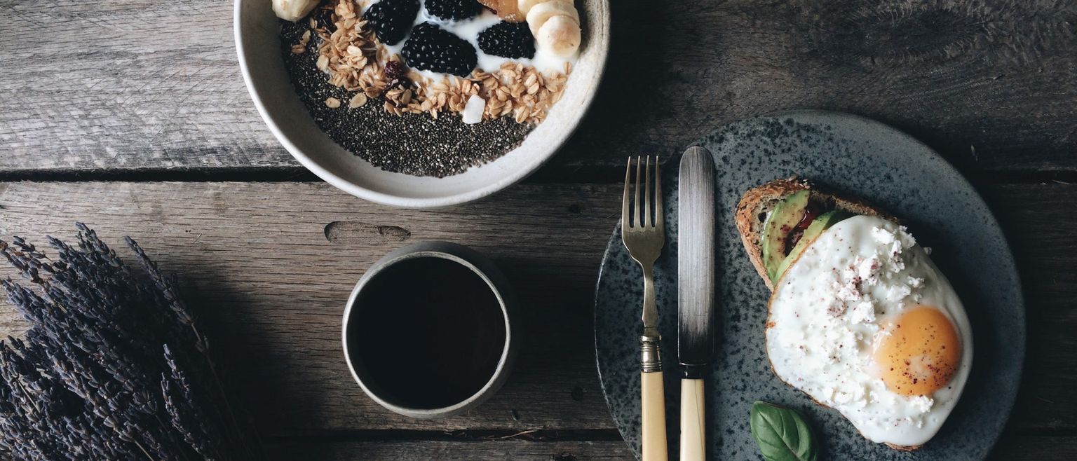 An aerial view of a healthy breakfast spread on a rustic wooden table. A bowl of chia pudding with granola, blackberries, and banana sits next to a cup of coffee and a plate with avocado toast topped with a fried egg.