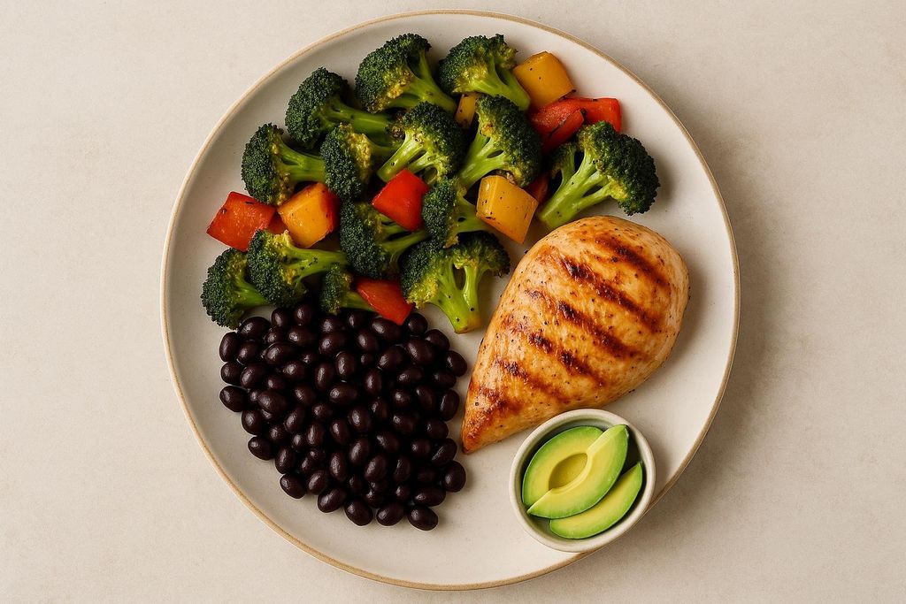A healthy meal on a white plate featuring a grilled chicken breast, steamed broccoli florets with red and yellow bell peppers, a mound of black beans, and a small dish with two slices of avocado.
