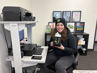 A woman sits at a desk with a computer and printer, holding a small black gift bag with green handles. She is smiling and wearing a black hoodie and beanie.
