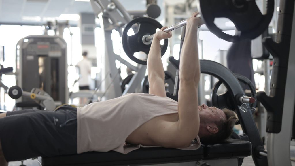A man is lying on a bench in a gym, lifting a barbell upwards during a bench press exercise.