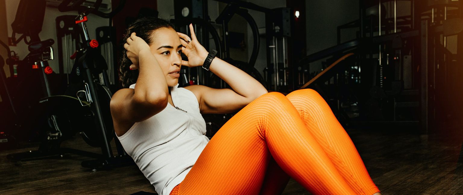 A woman in a white top and orange leggings exercises at the gym, doing crunches with her hands behind her head.