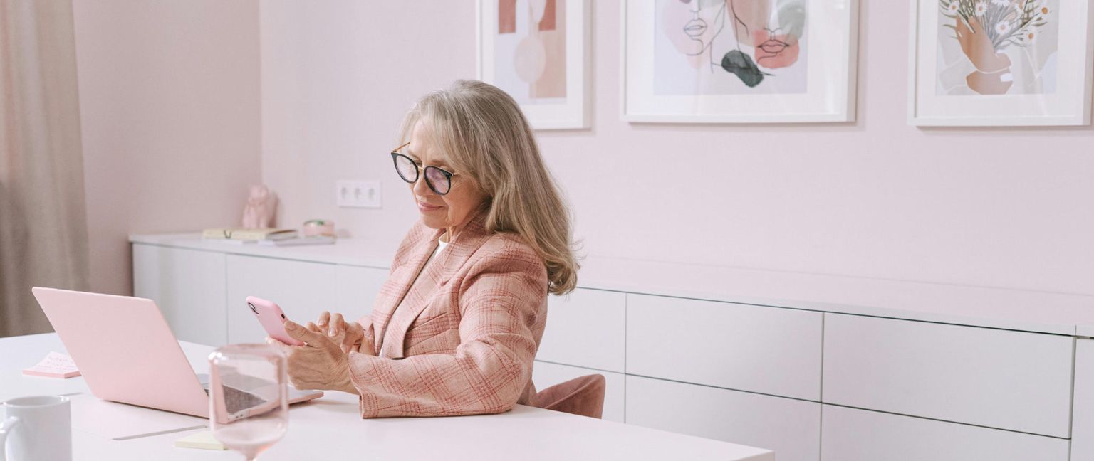 An older woman in a pink suit is sitting at a desk with a pink laptop, looking down at her pink smartphone and using the touchscreen.