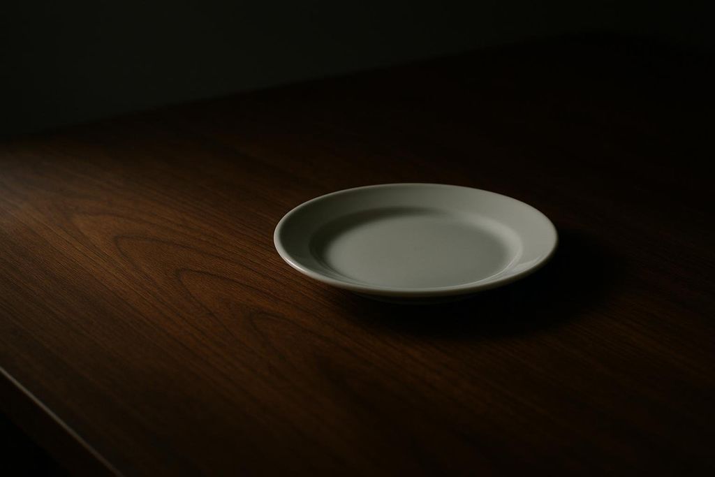 A light grey, empty dinner plate sits on a dark brown wooden table, partially illuminated by a soft light from the top left, casting most of the table into shadow.