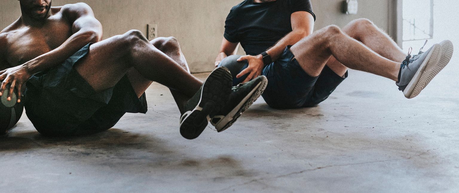 Two men performing medicine ball twists while sitting on the floor during a workout.