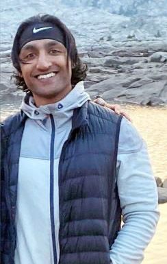 A man is smiling, wearing a black Nike headband, a light gray jacket with a hood, and a dark gray puffer vest.