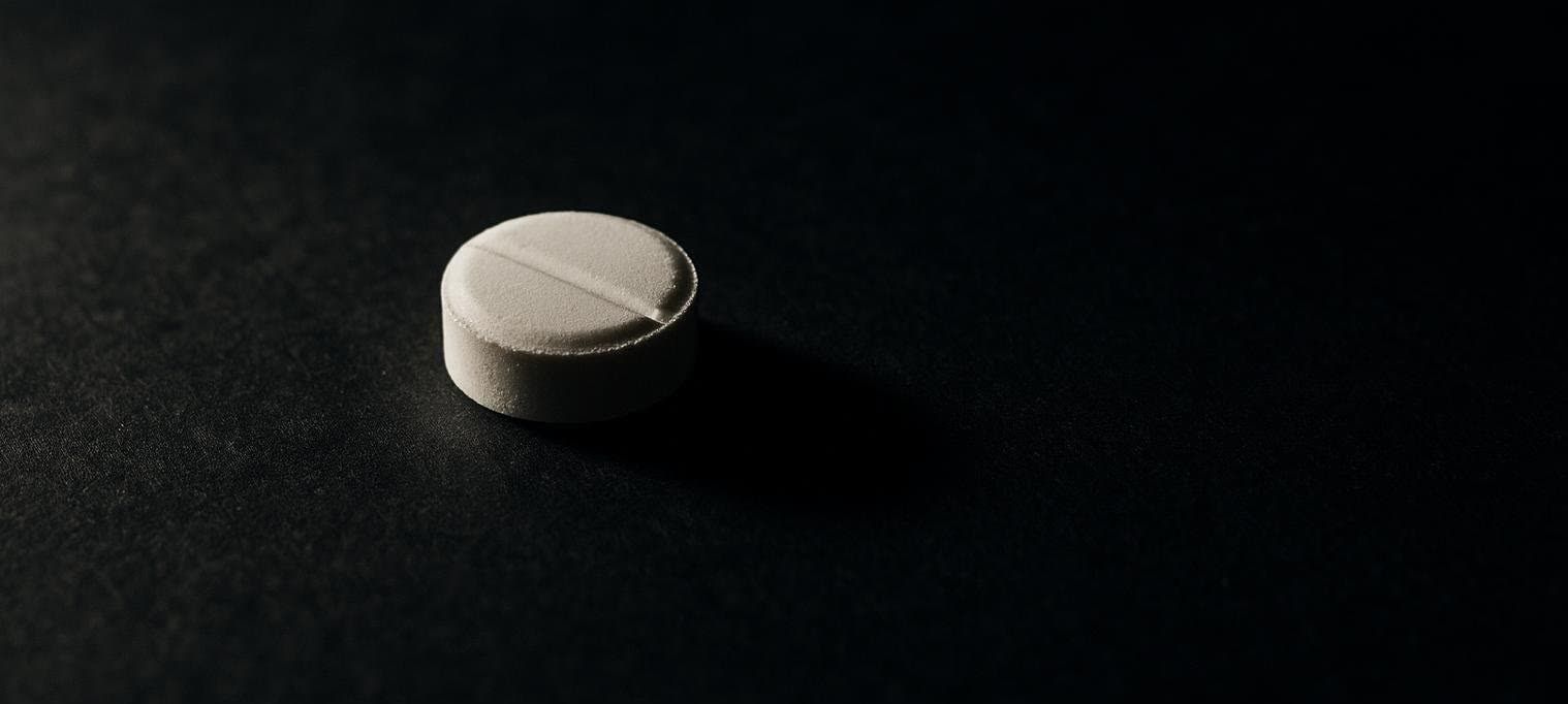 A close-up of a single white, round pill with a score mark, dramatically lit and casting a long shadow on a dark, textured surface.