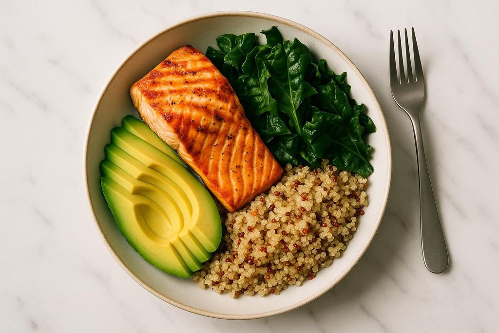 A close-up shot of a healthy bowl with a grilled salmon fillet, sliced avocado, green leafy vegetables, and quinoa, served on a white marble countertop with a fork beside it.