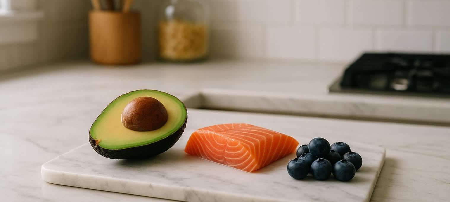 A close-up shot of a halved avocado, a raw salmon fillet, and a small pile of blueberries artfully arranged on a white marble cutting board. The background shows a blurry kitchen counter and stovetop.