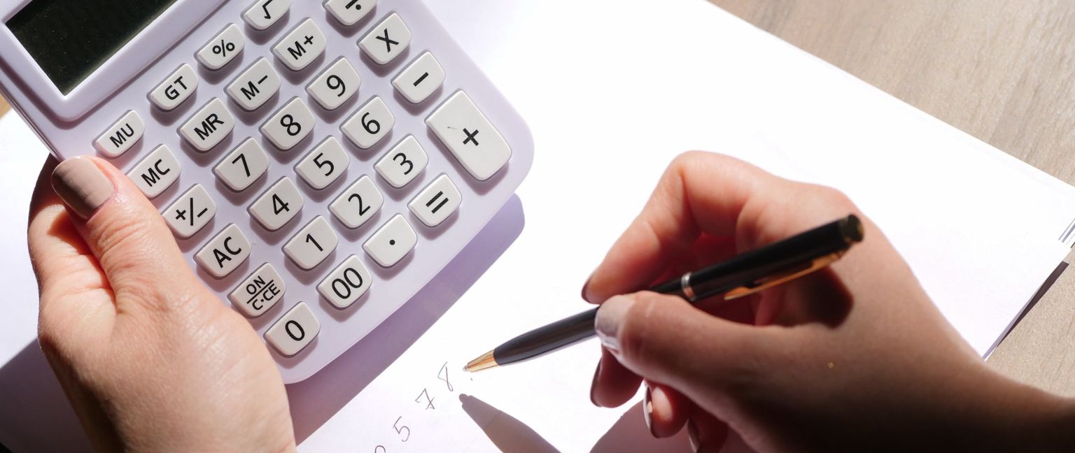 Close-up of someone's hands using a white calculator and writing numbers on a piece of paper with a black pen.