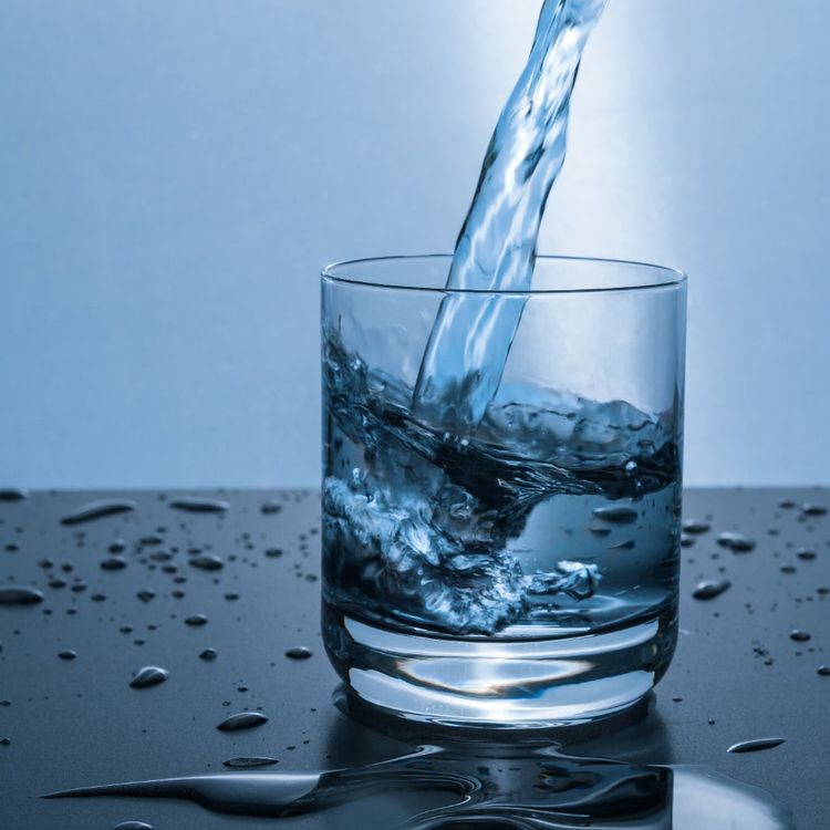 Water is being poured from above into a clear glass, splashing and filling the glass against a light blue background with water droplets on the surface below.