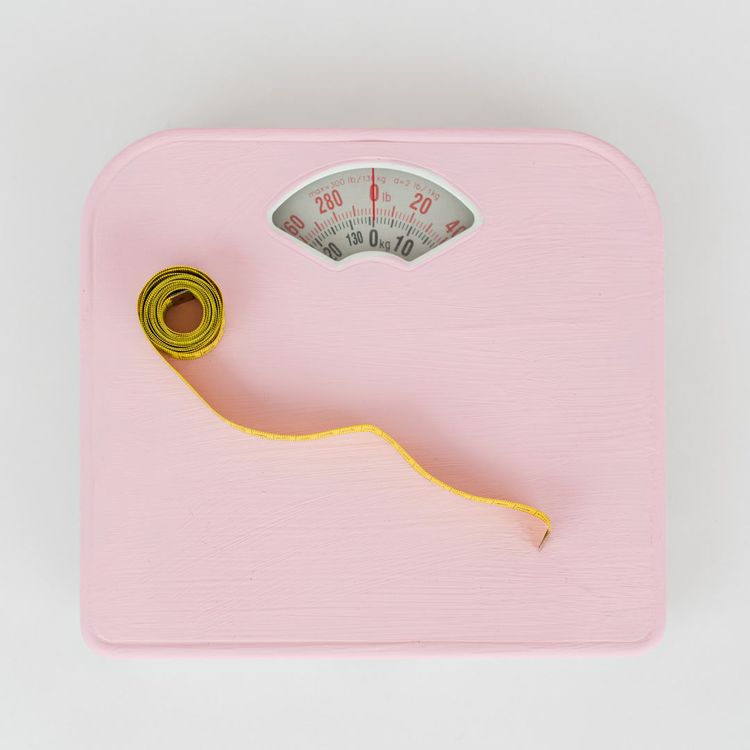 A pink bathroom scale with a coiled yellow measuring tape unspooling onto the scale. The scale reads '0 lb' and '0 kg'.