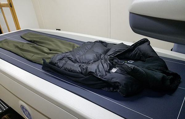 A black puffy vest and olive green athletic pants are laid out on the blue scanning bed of a medical imaging machine.