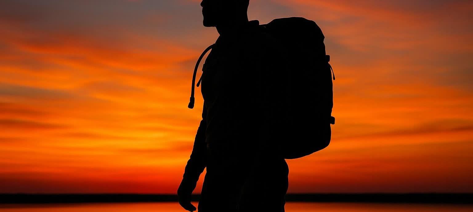 Silhouette of a person wearing a large backpack, or rucking, against a vibrant orange and red sunset over water.