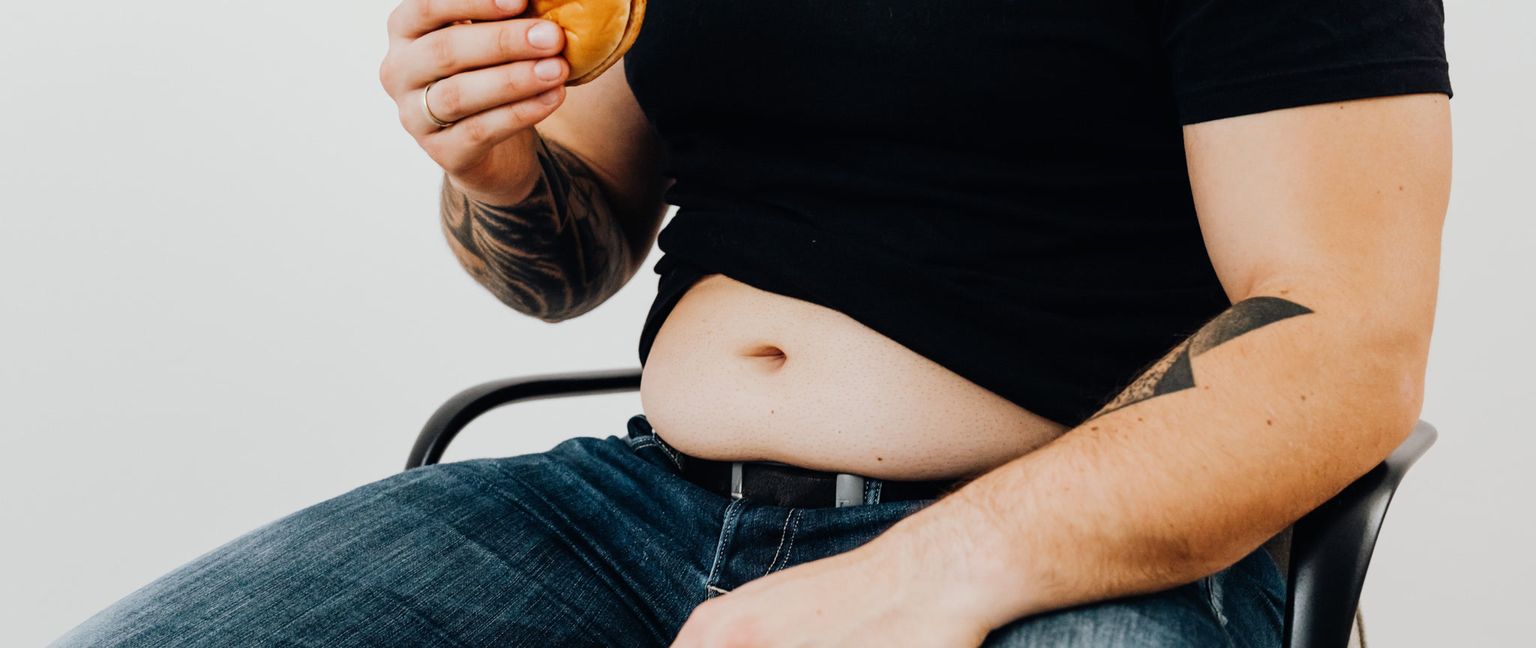 Close-up of a man holding a cheeseburger, revealing his belly over his jeans.