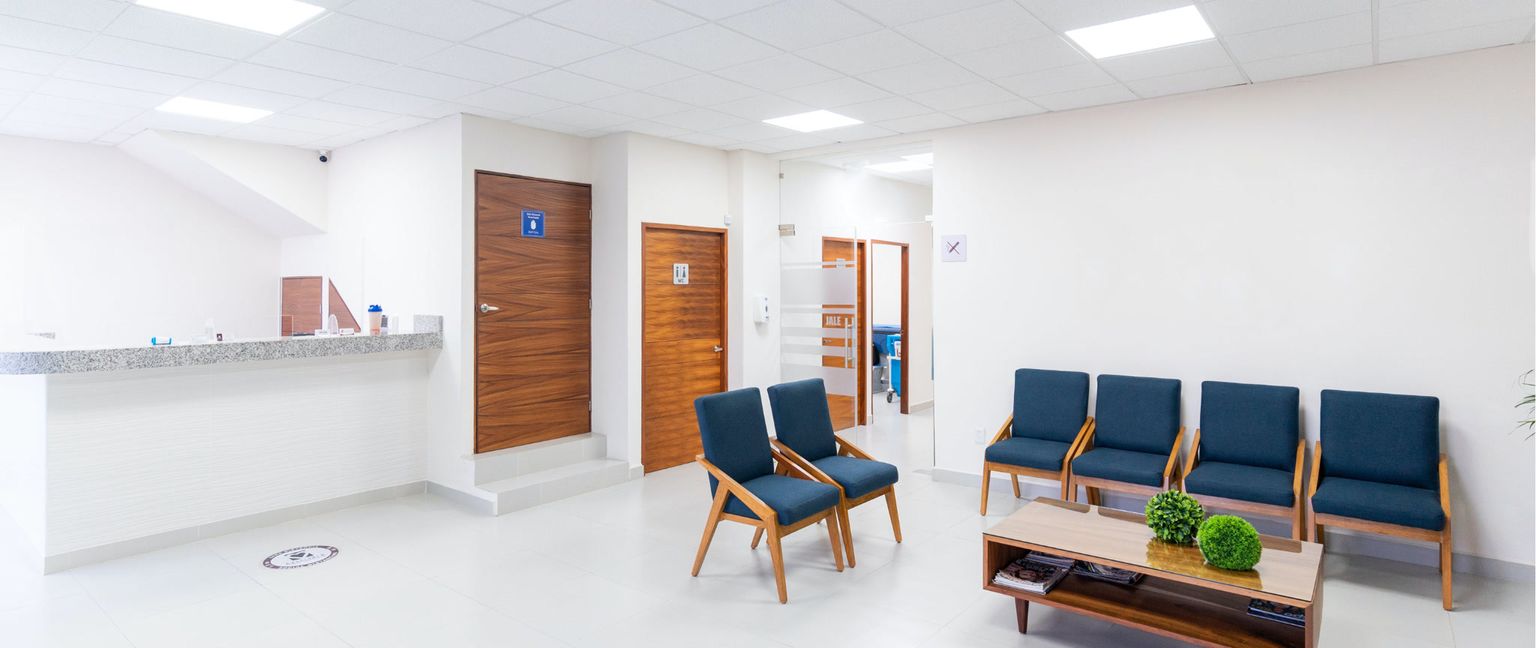A bright and modern medical waiting room with white walls, a granite reception desk, two large wooden doors, and several blue upholstered chairs around a coffee table.
