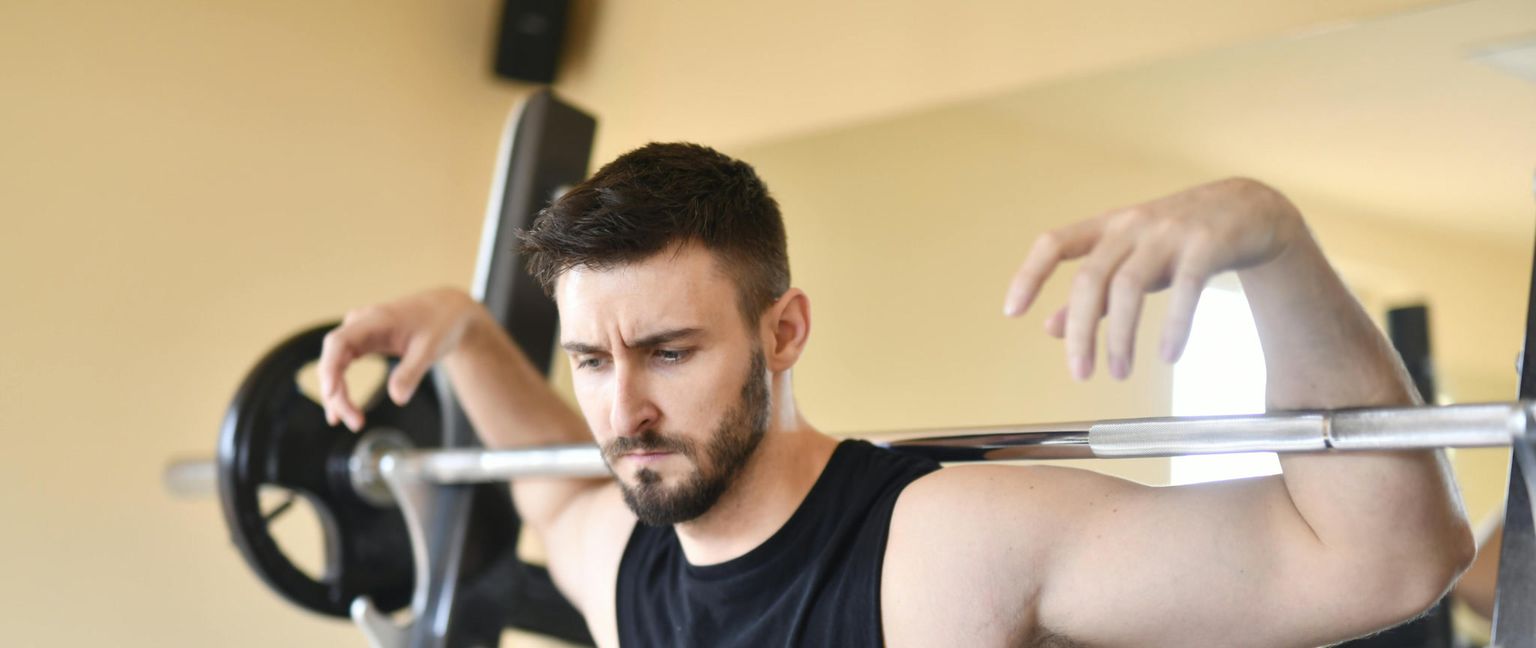 A man with a beard in a black tank top with a barbell on his shoulders, preparing to squat in a gym.