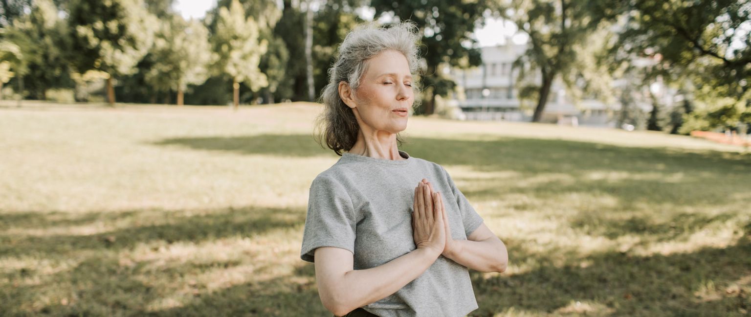 An older woman with gray hair stands in a park and meditates with her eyes closed and hands pressed together in prayer position. The sun shines through the trees, casting dappled shadows on the grass.
