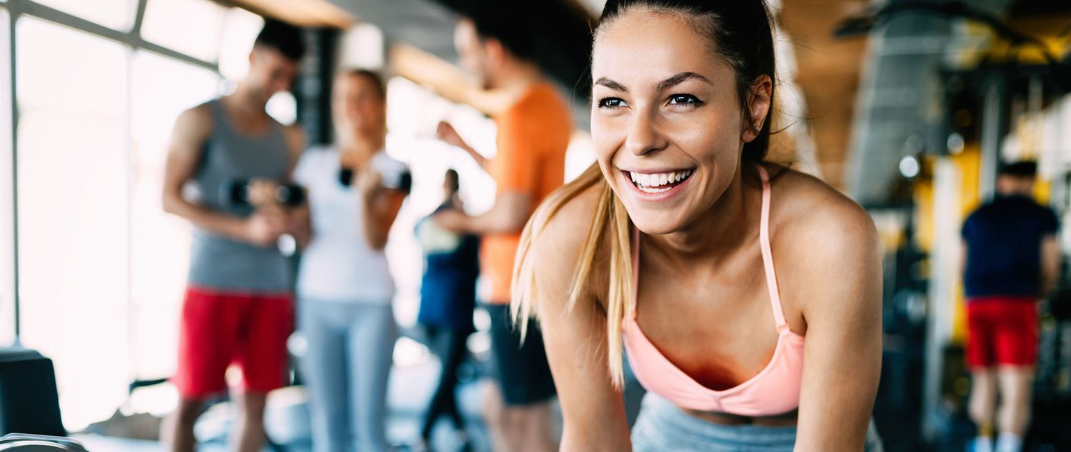 A young woman smiles brightly while exercising at a gym, with other people working out in the background.