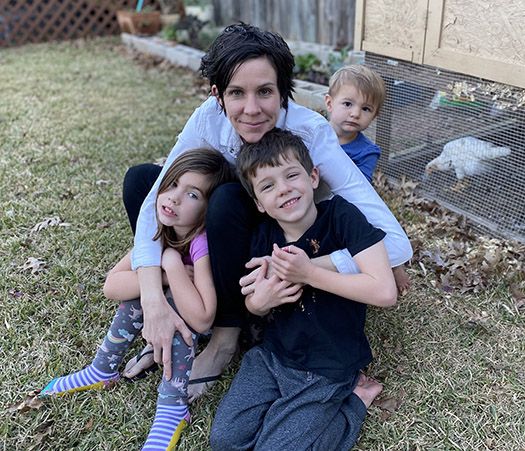 A woman with dark, short hair is sitting on grass and smiling. She is holding a young girl wearing socks with unicorns and rainbows and a young boy wearing a black t-shirt and grey sweatpants. Another young boy is sitting behind the woman's right shoulder, looking off to the side. In the background is a wooden chicken coop with a chicken visible inside.