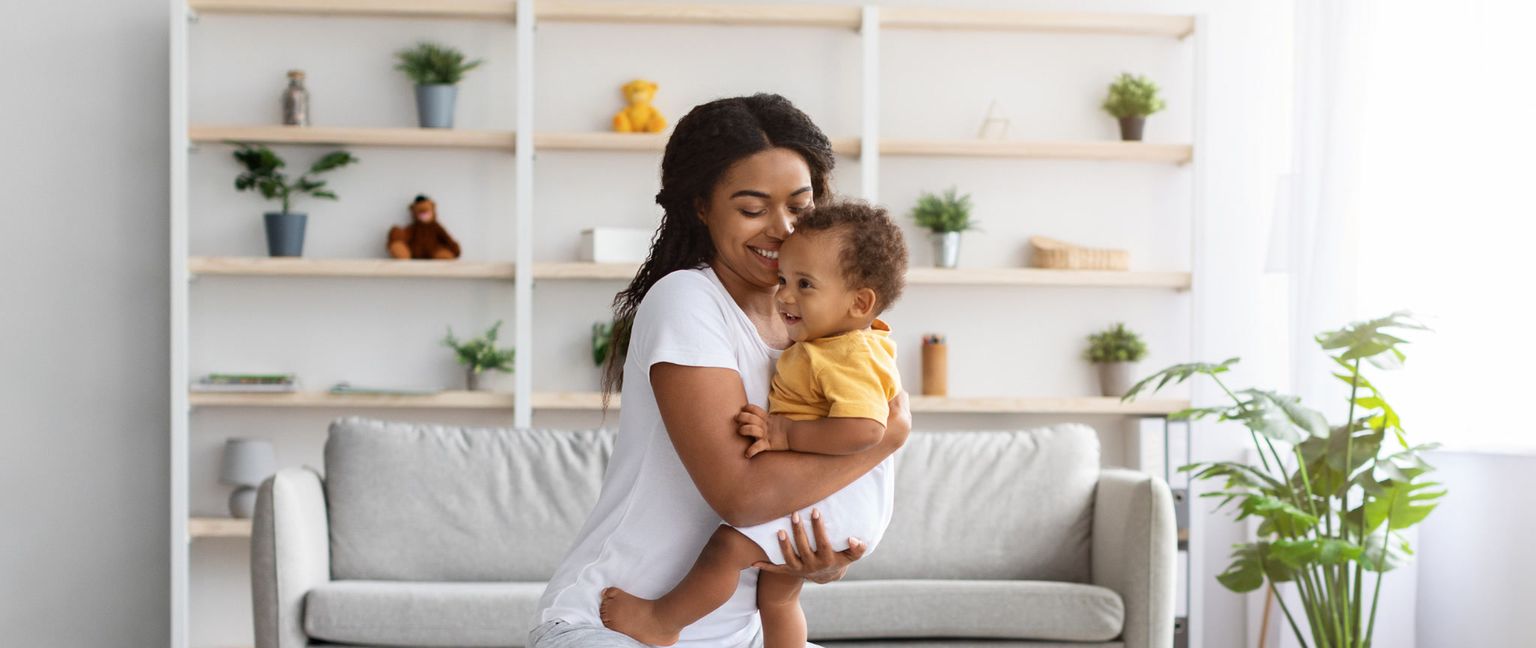 A smiling African American mother holds her happy baby in a bright living room.