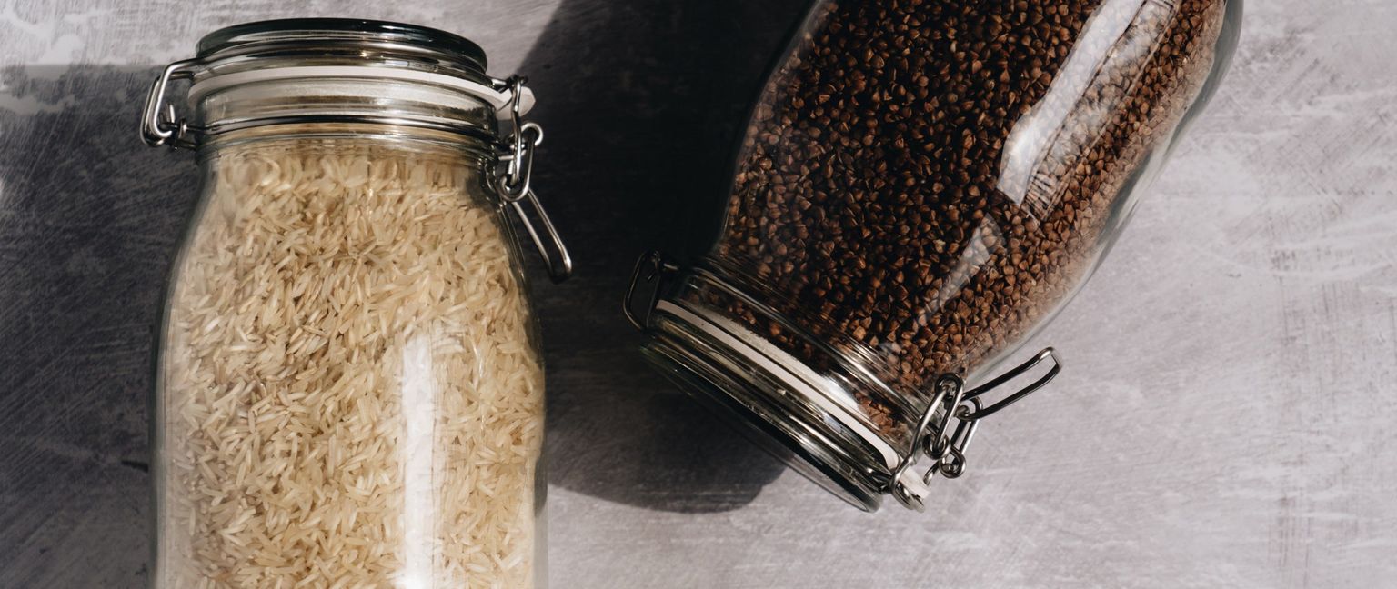Two jars filled with different types of grain. The jar on the left contains long-grain rice, and the jar on the right contains buckwheat.