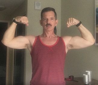 A man wearing a red tank top flexes his bicep muscles with his arms held up and out to his sides.