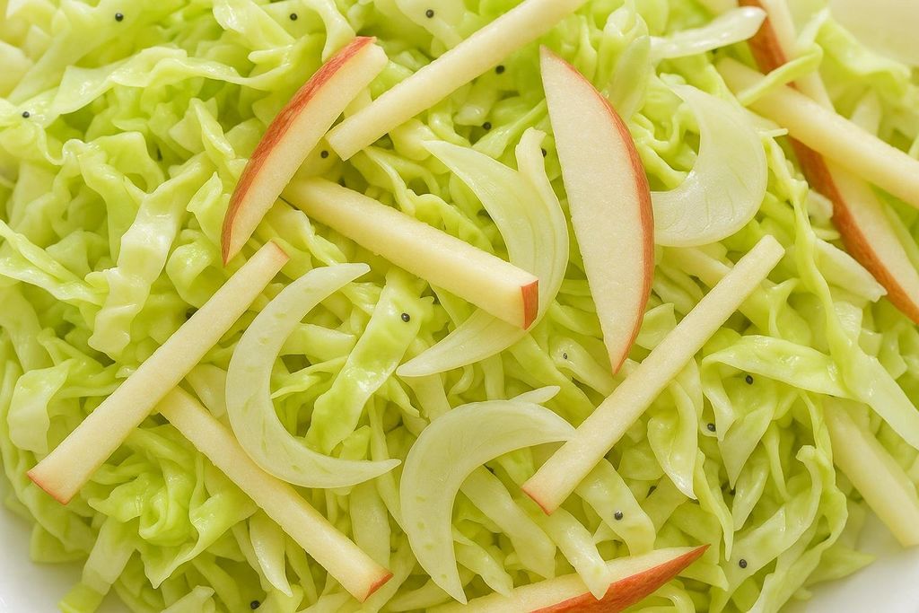 A close-up shot of an apple-fennel slaw made with thinly shredded green cabbage, sliced fennel, and red-skinned apple sticks, scattered with a few black pepper flakes.