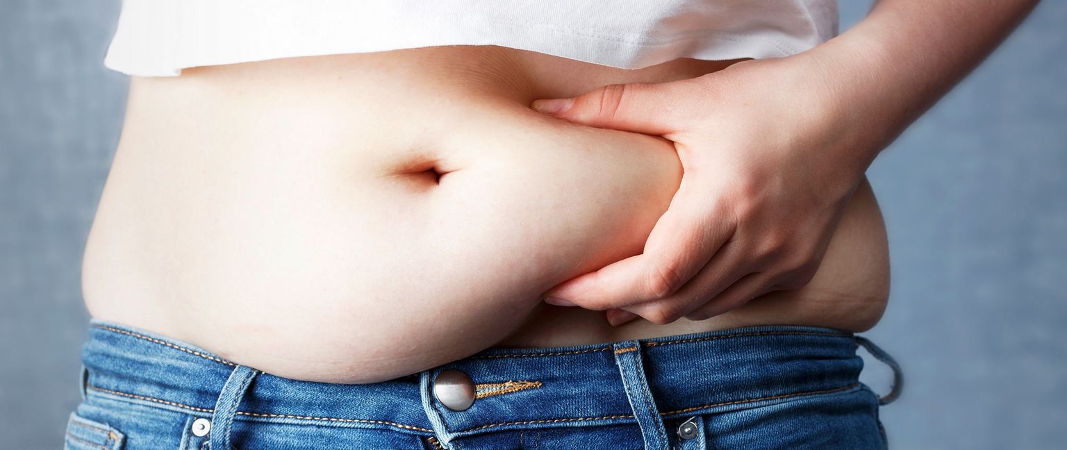 A person's hand pinches a roll of fat on their belly. They are wearing a white shirt and blue jeans.