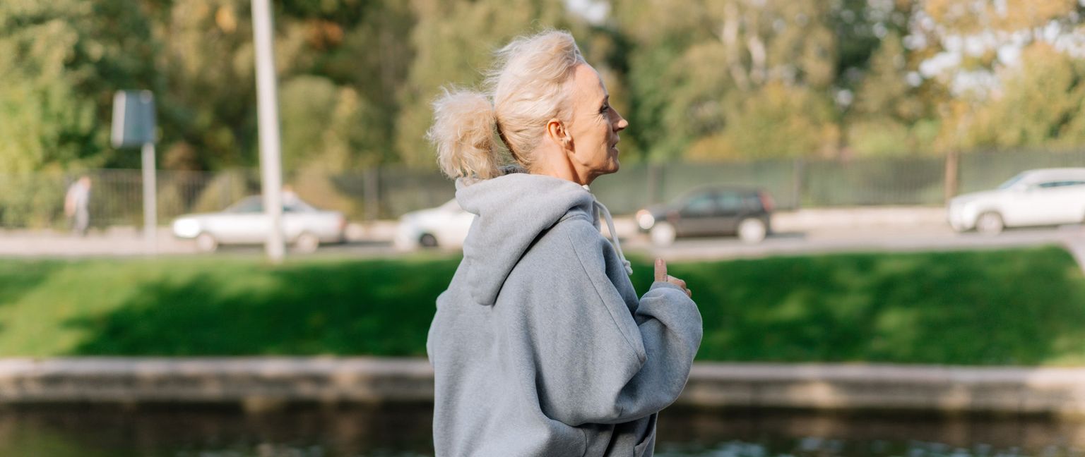 A side profile view of an older woman with blonde hair tied up in a ponytail, wearing a grey hoodie, jogging outdoors.