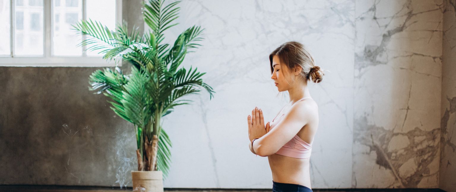 A young woman meditating with her eyes closed and hands clasped in front of her chest. A plant and smoking incense are visible to her left.