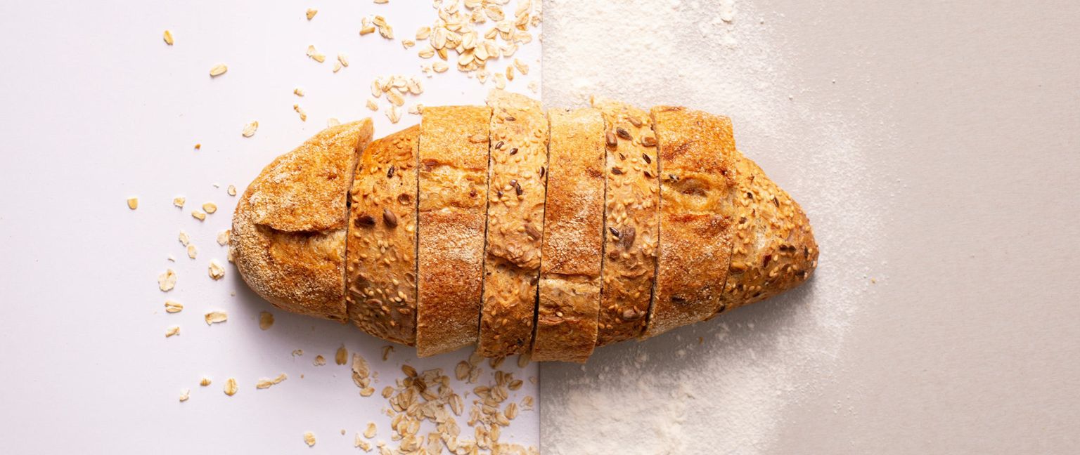 A sliced loaf of bread, rich with visible grains, rests across a surface split down the middle. The left side of the surface is white with scattered oatmeal, while the right side is tan dusted with flour.