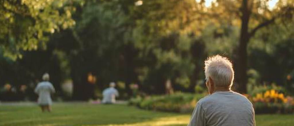 An elderly man sits in a park, with blurred figures of other people in the background, enjoying the late afternoon sun filtering through the trees.