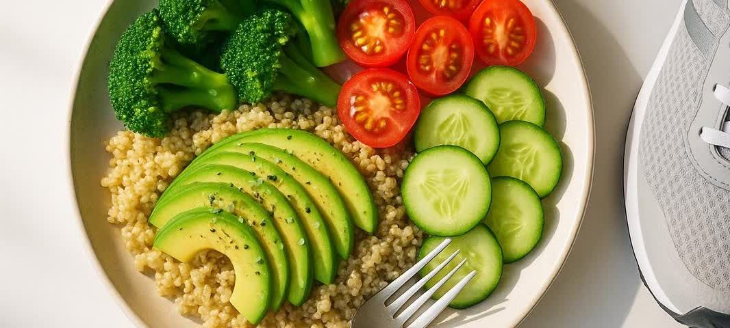 A white plate holds a healthy meal of quinoa, sliced avocado, broccoli, halved cherry tomatoes, and sliced cucumbers. Next to the plate, the toe of a grey running shoe is visible, suggesting a connection between diet and exercise.