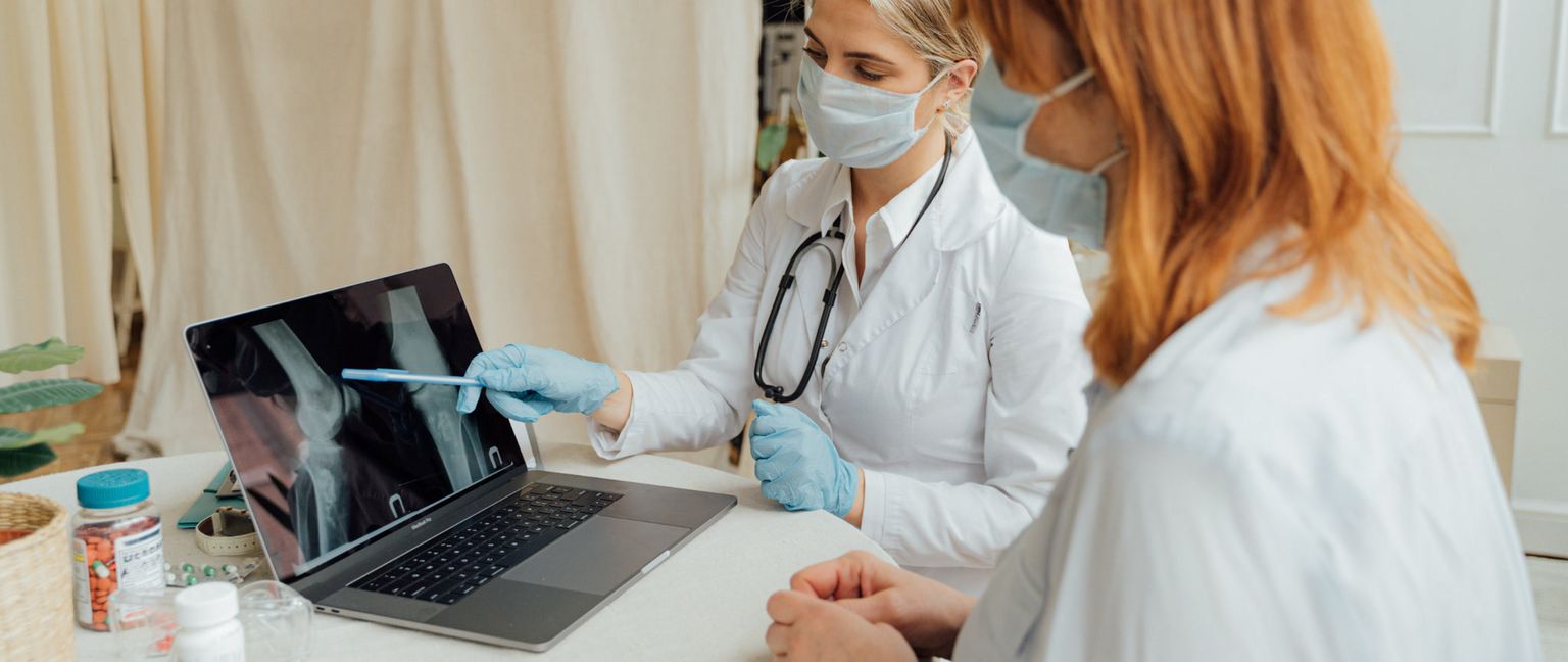 A female doctor in a lab coat and mask points to a bone scan on a laptop while a patient in a white shirt and mask listens.