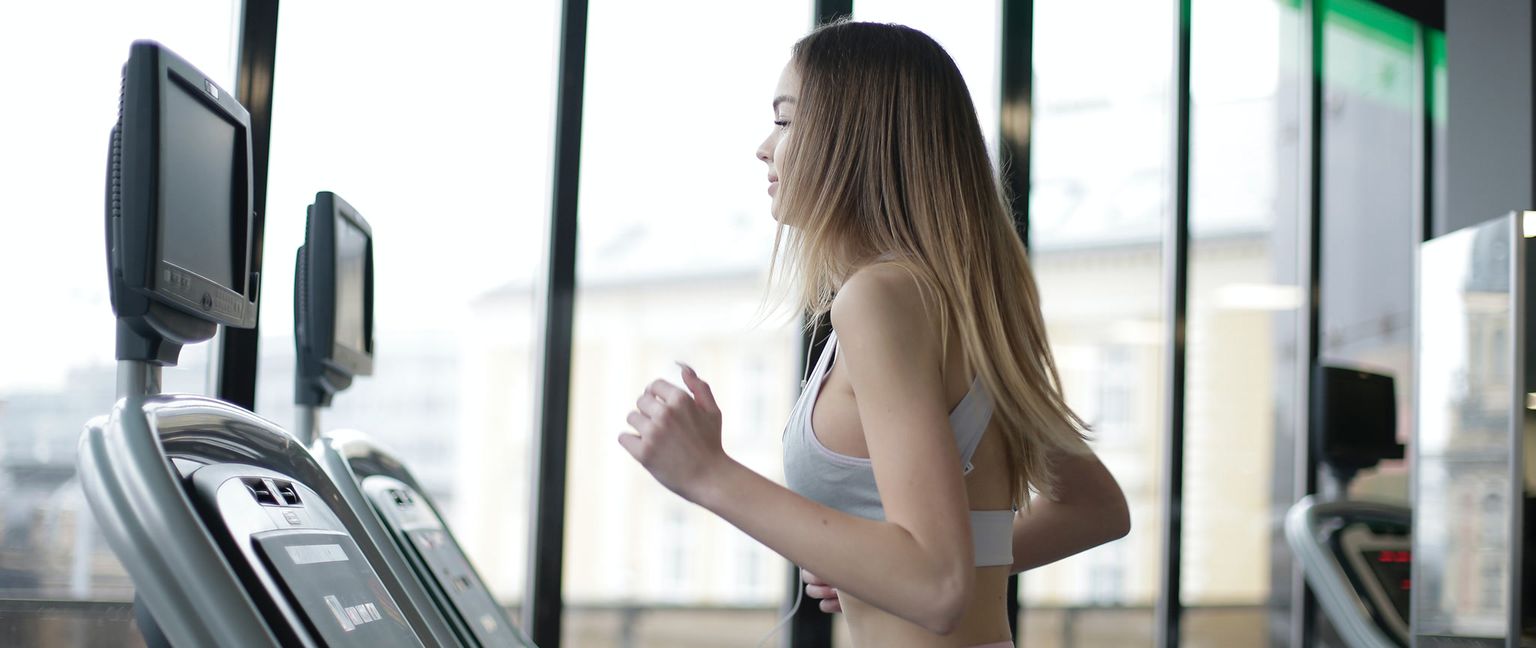 A woman with blonde hair running on a treadmill in a gym with large windows.