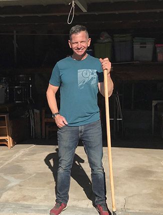 A man in a blue shirt and jeans stands holding a long stick in a garage.
