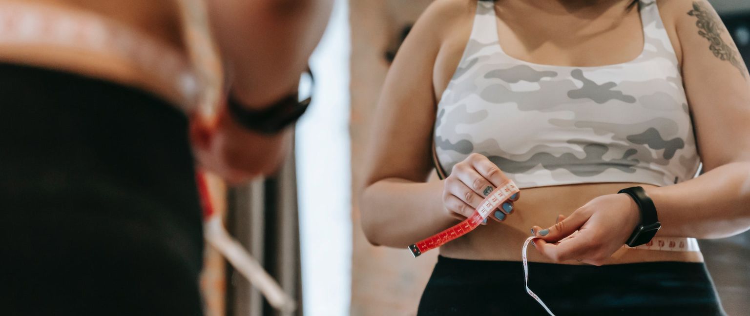 A woman in a tank top measures her waist with a red and white measuring tape, looking in a mirror.