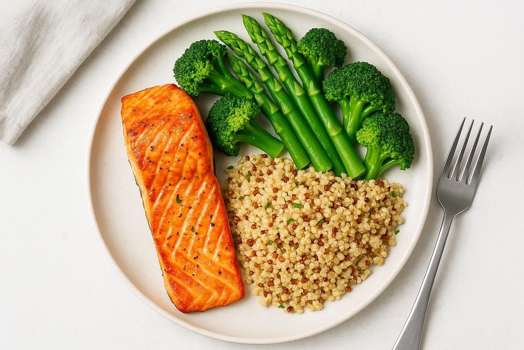 A healthy meal with a salmon fillet, a serving of quinoa, and green vegetables including broccoli florets and asparagus spears, presented on a white plate with a fork nearby.