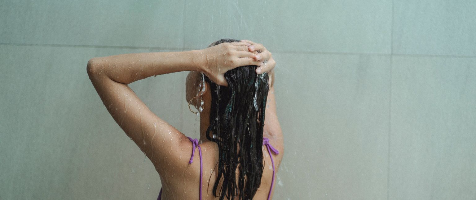 Rear view of a woman washing her dark hair under running water in a shower.