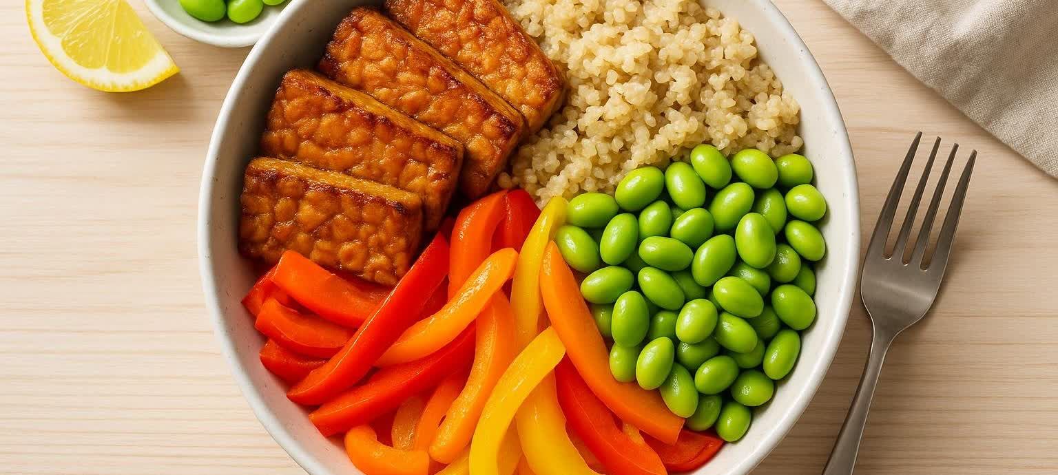A top-down view of a colorful and appetizing high-protein vegan power bowl composed of brown tempeh slices, light-brown quinoa, bright green edamame, and vibrant red and yellow bell pepper strips, all served in a white bowl on a light wooden surface. A fork and a lemon wedge are visible beside the bowl.