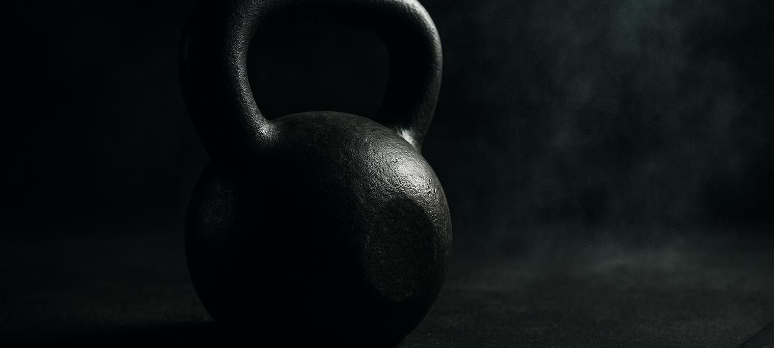 Close-up photo of a black kettlebell on a dark gym floor, with visible chalk dust particles in the air, creating a dramatic effect.