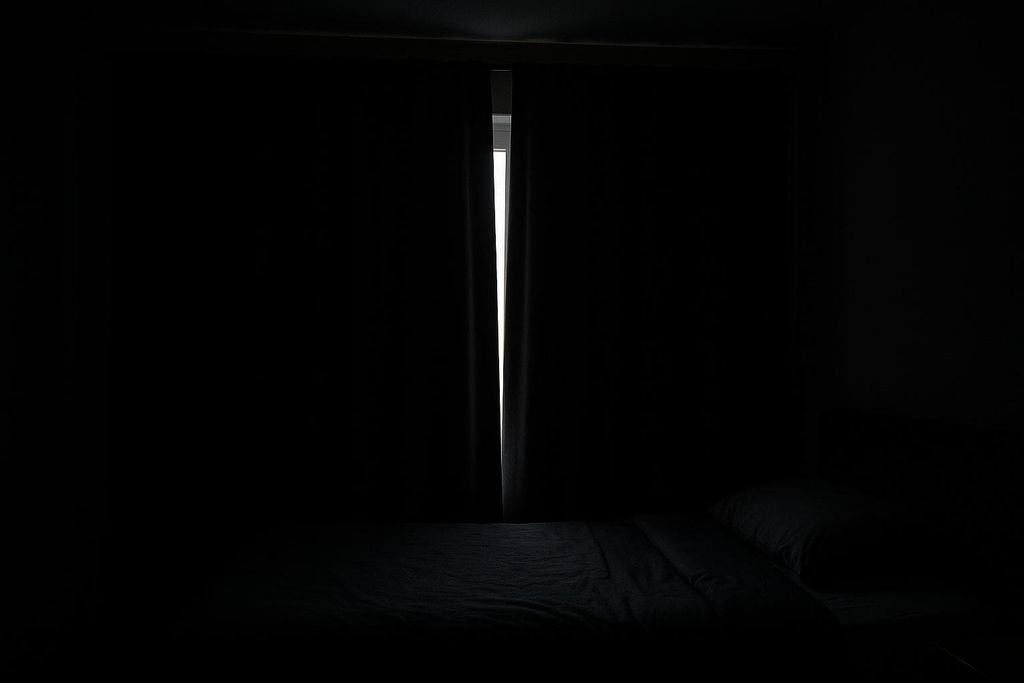 A very dark bedroom interior with blackout curtains drawn, leaving just a sliver of bright light showing through the gap in the middle, revealing a bed with dark bedding in the foreground.