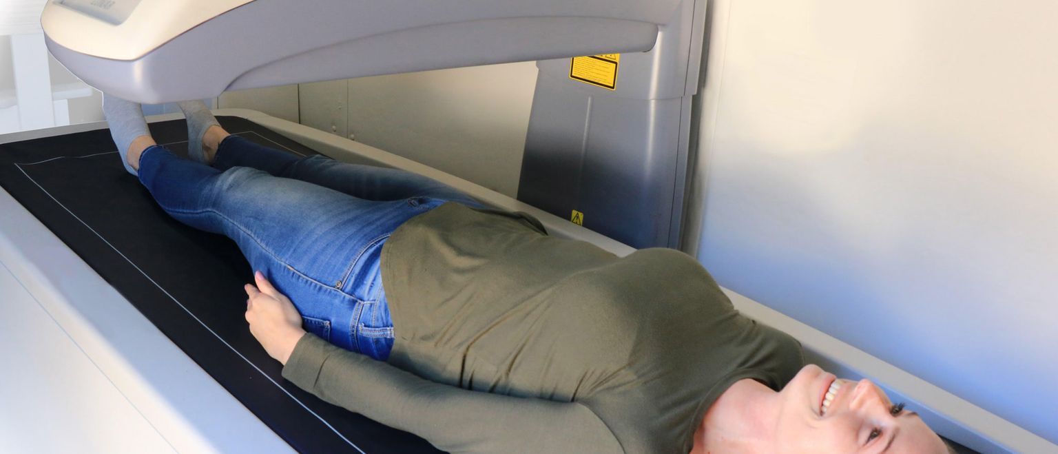 A woman in jeans and a green shirt lies smiling on a medical scanning machine.