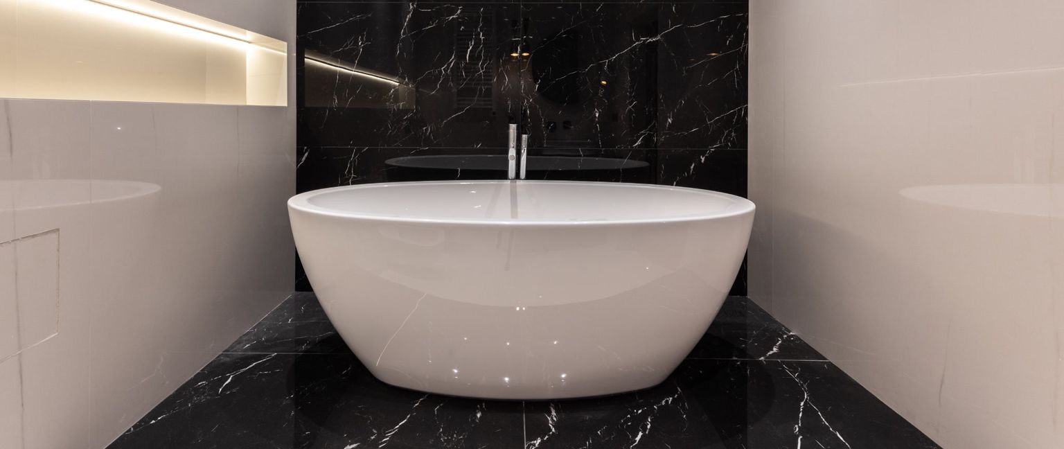 A wide white, freestanding bathtub sits in a modern bathroom with black marble floor and wall tiles and white wall tiles. A silver faucet stands behind the tub.