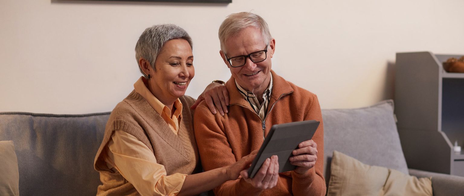 A senior couple sitting on a sofa, smiling and looking at a digital tablet.