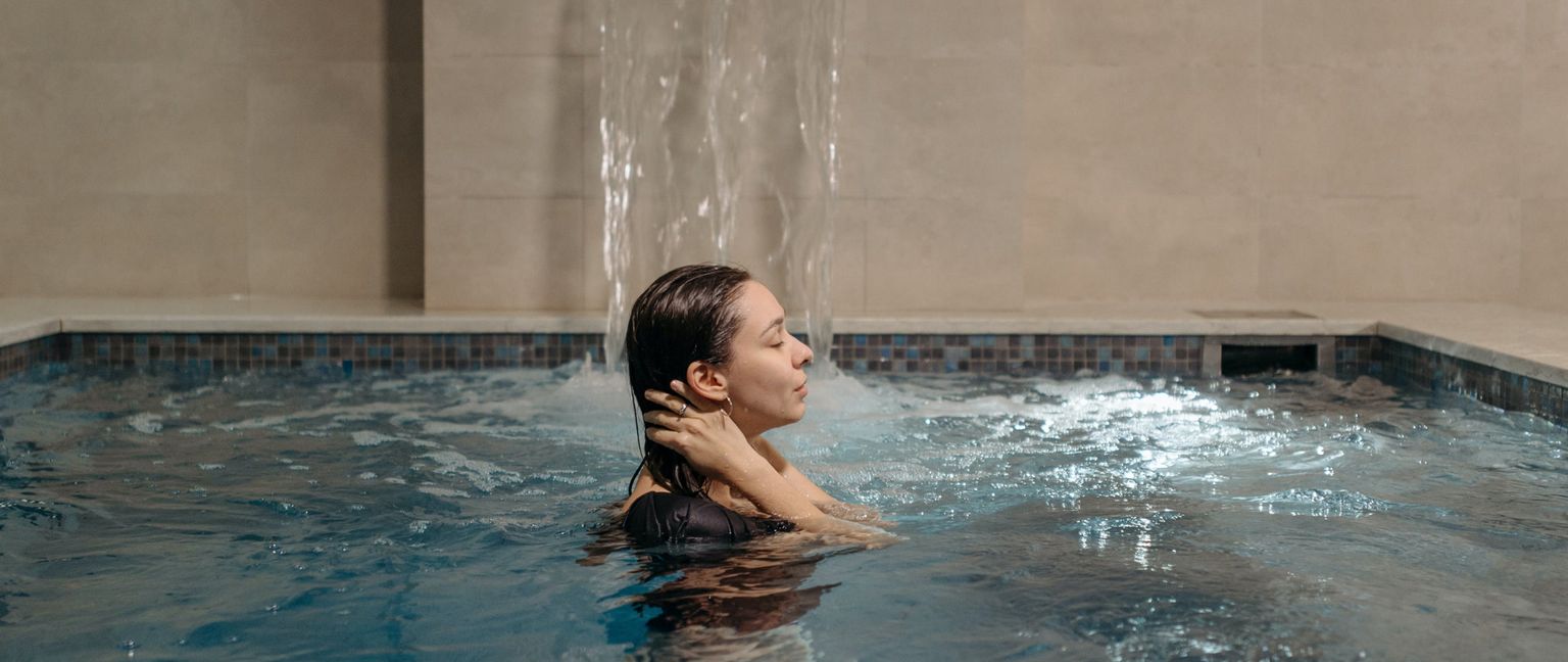 A woman with her eyes closed, relaxing in an indoor pool with water cascading behind her from a spout.