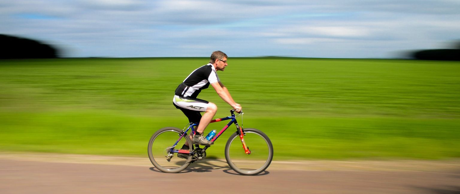 A man wears cycling gear and glasses while riding a mountain bike along a road parallel to a vibrant green field under a partly cloudy sky. The image has a motion blur effect, suggesting speed.
