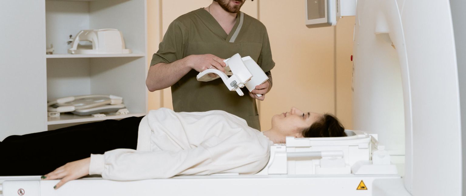 A patient lying on a CT scanner bed while a medical technician holds a head restraint.